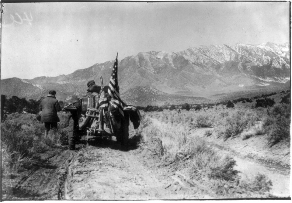 New York to Paris auto race, 1908- rear view of auto with tire chains and U.S. flag on crude road; high mountains in distance LCCN2004679575