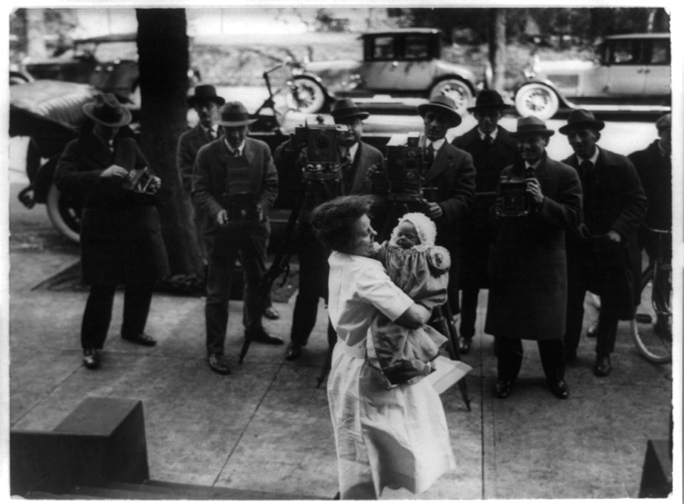 News photographers photographing Paulina Longworth, daughter of Nick and Alice Longworth, being held by woman on sidewalk, Washington, D.C. LCCN89713381
