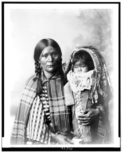 Pee-a-rat and baby - Utes. Half-length portrait of woman, facing front, with plaid blanket on and checked shirt, holding baby in cradleboard LCCN94509883