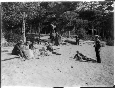 People relaxing on sandy beach in the Adirondack Mts., N.Y.; lean-tos in backgrd LCCN2003688923