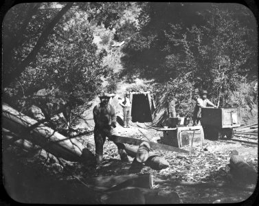 Photograph of four men outside a mine shaft. The foremost man appears to be cutting timbers for bracing the interior of the shaft. (5285433767)