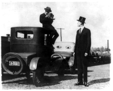 Photographer seated on top of car photographing tall man in top hat LCCN92501040