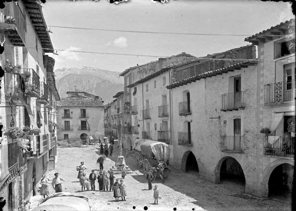 Plaça major porxada amb gent, carros i una creu al centre al poble de Campo
