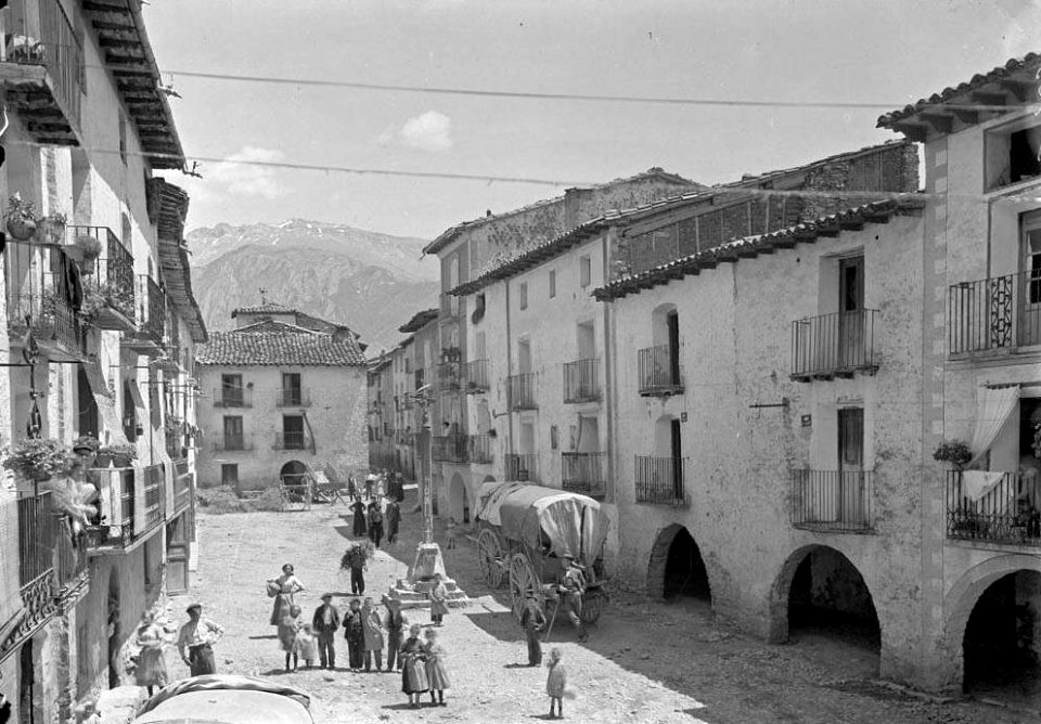 Plaça major porxada amb gent, carros i una creu al centre al poble de Campo (cropped)