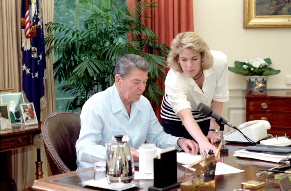 President Ronald Reagan sitting at his desk prior to his radio address to the Nation on International Trade in the Oval Office with Caroline Rhoden