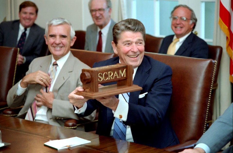 President Ronald Reagan sitting next to Senator Paul Laxalt holding up a scram sign during a Cabinet Room meeting