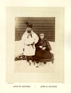 Samisk par fra Kautokeino sitter på baksiden av en pulk. De er kledd i pesk, kofte, sjal og skaller - Norsk folkemuseum - NF.15006-075