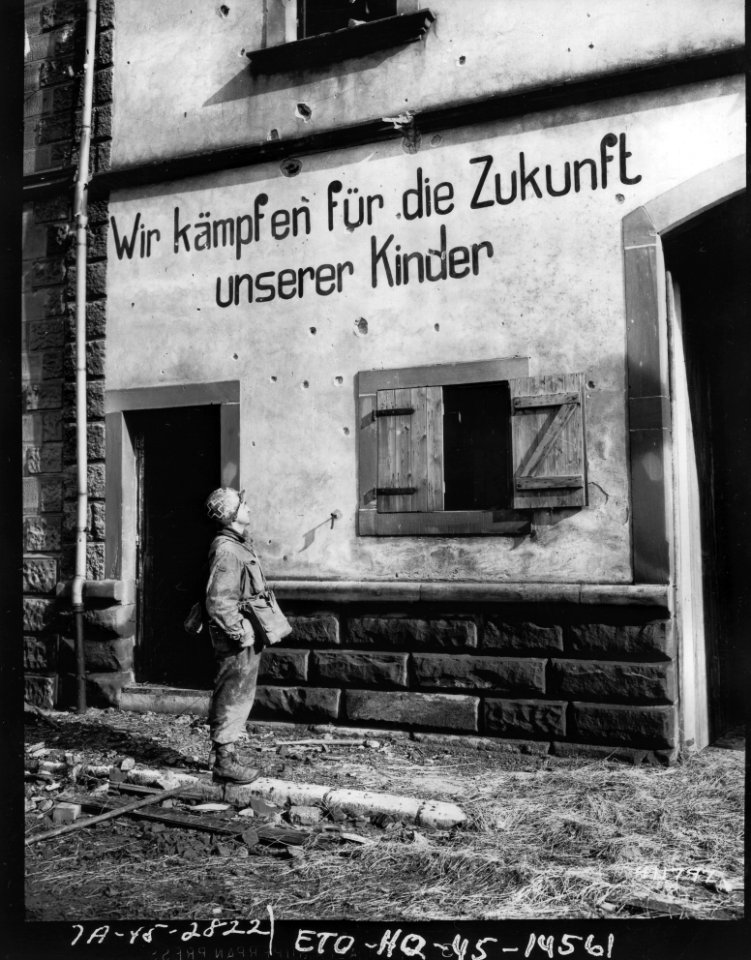 SC 411797 - PFC Abraham Green, medic, New Haven, Conn., 253rd Regiment, 63rd Division, stops, reads and wonders at a German sign, We fight for the future of our children. 21 February, 1945