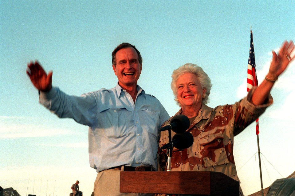 President George H. W. Bush and First Lady Barbara Bush wave as they stand in the back of a vehicle during a visit to a desert encampment