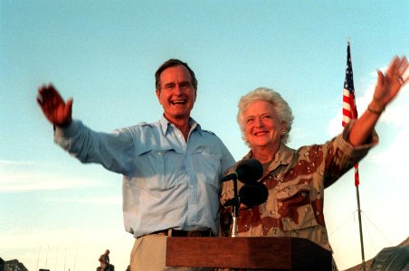 President George H. W. Bush and First Lady Barbara Bush wave as they stand in the back of a vehicle during a visit to a desert encampment