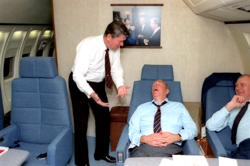 President Ronald Reagan joking with a sleeping Secretary of State George Shultz aboard Air Force One during a trip to South America