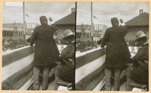 Theodore Roosevelt on flag-draped platform, with his back to the camera, speaking to the crowd LCCN2013651072