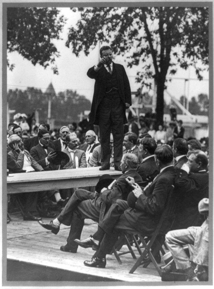 Theodore Roosevelt standing on table outdoors and making speech to men seated around him, right hand raised) - Smith Art Photography, Freeport, Ill LCCN91787238
