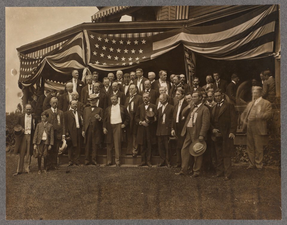 Theodore Roosevelt standing in center of a group of men outside a porch draped with American flags LCCN2013650557