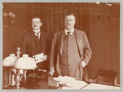 Theodore Roosevelt standing behind desk, facing slightly left, with Presidential Secretary William Loeb LCCN2009631477