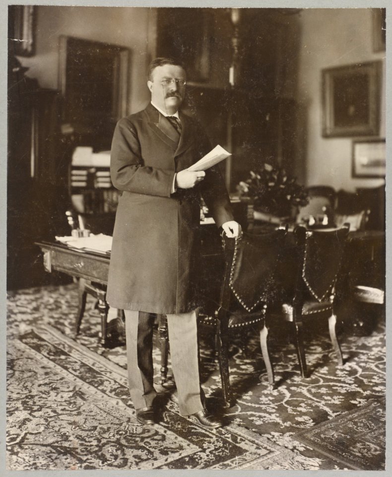 Theodore Roosevelt, full-length portrait, standing by table, facing right; holding paper in right hand, left hand on back of chair LCCN97502260