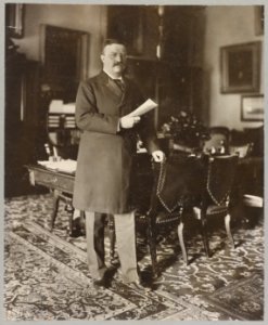 Theodore Roosevelt, full-length portrait, standing by table, facing right; holding paper in right hand, left hand on back of chair LCCN97502260