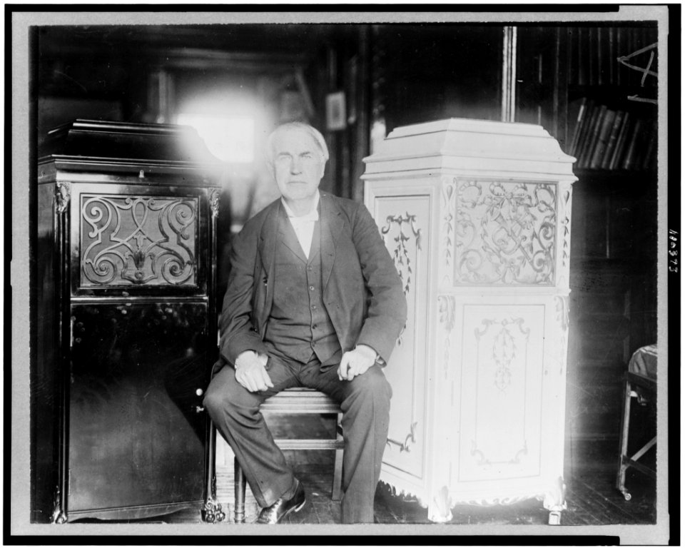 Thomas Alva Edison, full-length portrait, seated, facing front, between two phonograph cabinets, one of which is made of concrete LCCN94504410