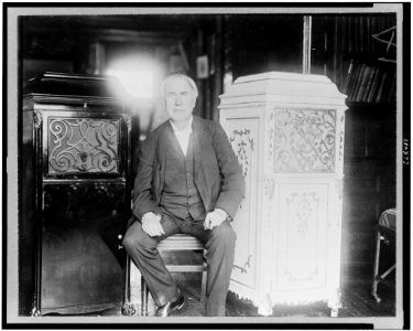 Thomas Alva Edison, full-length portrait, seated, facing front, between two phonograph cabinets, one of which is made of concrete LCCN94504410