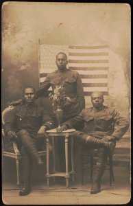 Three unidentified African American soldiers in uniforms with vase of flowers in front of American flag and painted backdrop LCCN2017648686 photo