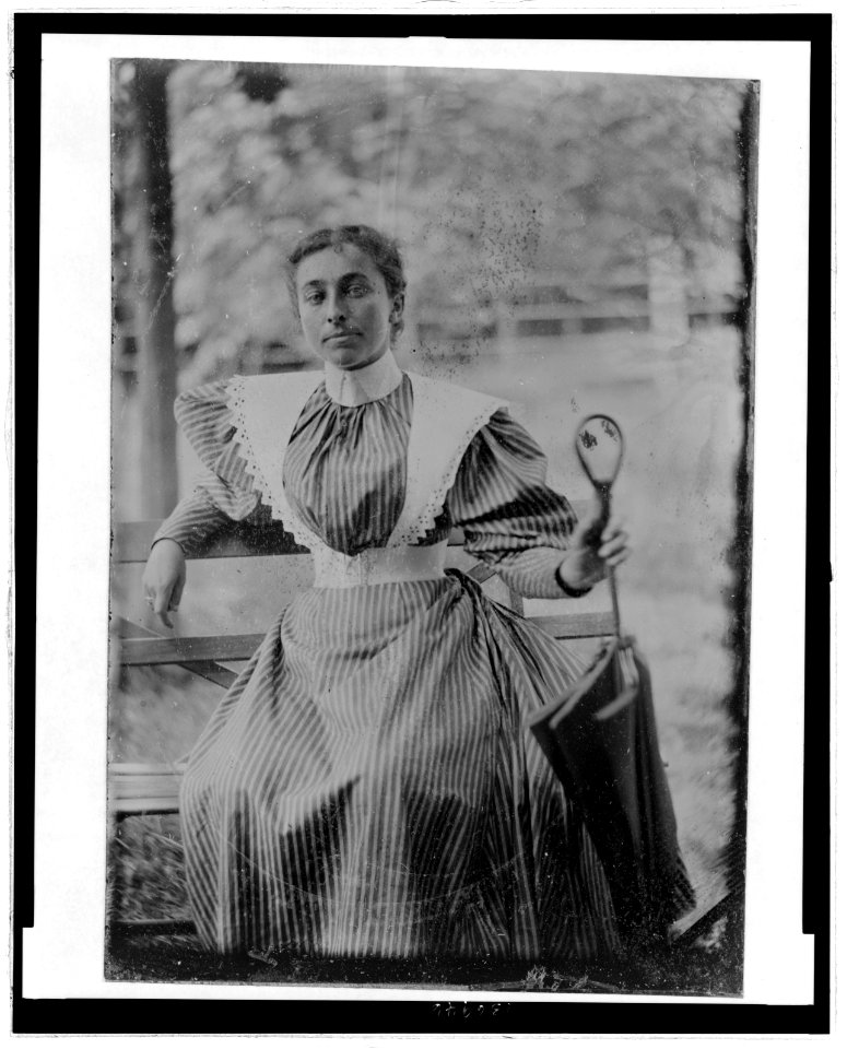 Three-quarter length portrait of a young woman wearing a dress with mutton-chop sleeves and high collar, seated holding an umbrella LCCN2006689568