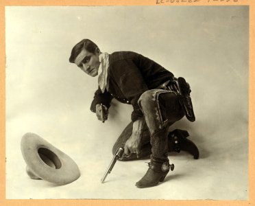 Tom Mix, full-length portrait, kneeling, facing left, holding two guns, hat on floor, in Mr. Logan, U.S.A. LCCN96508056