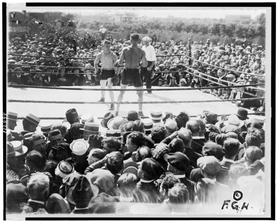 Two boxers and referee in outdoor boxing ring, surrounded by audience LCCN99402329