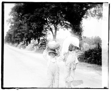 Two little boys in overalls standing in a dirt road, balancing loads on their heads LCCN2016828300