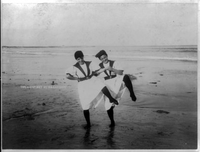 Two girls standing together on beach, each with their left foot raised LCCN2003688917 photo