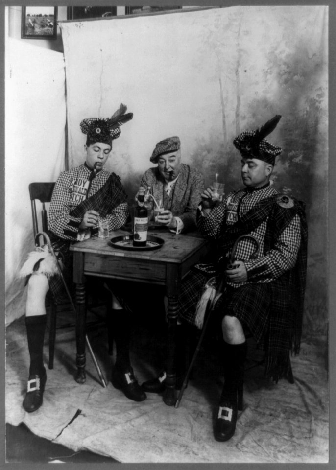 Two men wearing kilts and man smoking cigar, seated at small table drinking Sandy MacDonald scotch LCCN89706570