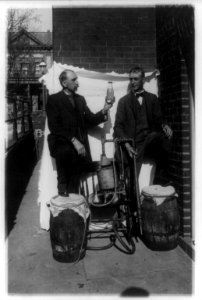 Two men standing outdoors with small still, one of them holding up bottle of liquor LCCN88715935