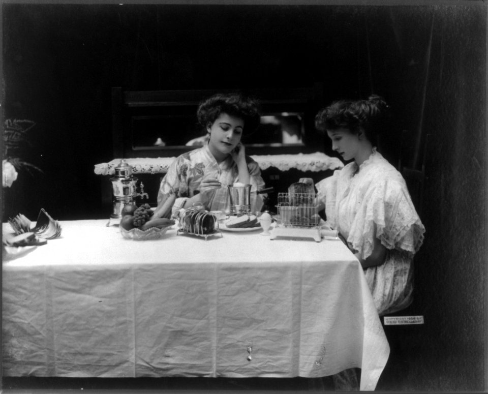 Two women seated at breakfast table with electric heating and cooking appliances LCCN2005685987
