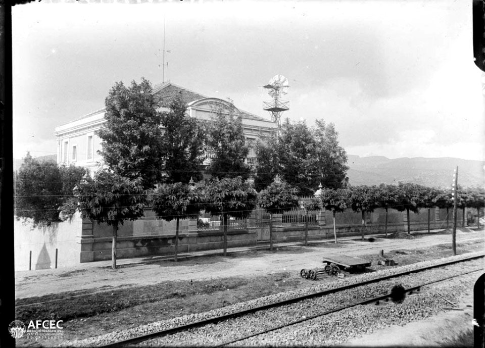 Un edifici de la Garriga amb uns arbres davant i les vies del tren en primer terme
