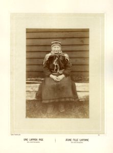 Ung samisk kvinne med kofte og sjal sitter på en benk utenfor bygning - Norsk folkemuseum - NF.15006-038