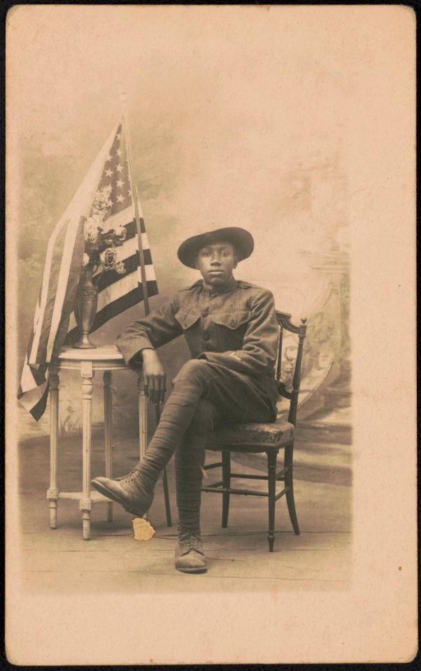 Unidentified African American soldier in uniform and campaign hat sitting next to vase of flowers and American flag in front of painted backdrop LCCN2017648682