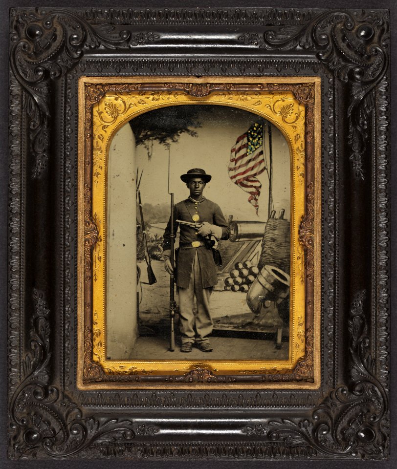 Unidentified African American soldier in Union uniform with a rifle and revolver in front of painted backdrop showing weapons and American flag at Benton Barracks, Saint Louis, Missouri LCCN2010647218