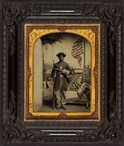 Unidentified African American soldier in Union uniform with a rifle and revolver in front of painted backdrop showing weapons and American flag at Benton Barracks, Saint Louis, Missouri LCCN2010647218