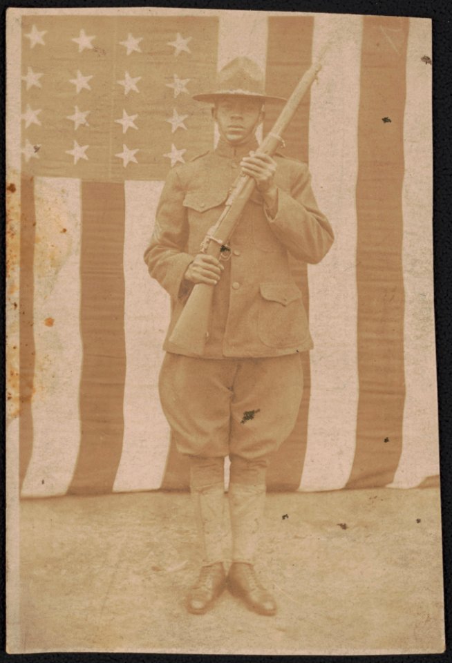 Unidentified African American soldier in uniform and campaign hat with rifle in front of American flag LCCN2017648689