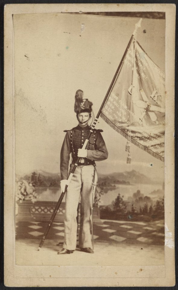 Unidentified soldier in artillery uniform and Shako hat with Battery K, 1st U.S. Artillery flag and battle honors in front of painted backdrop showing garden leading to river) - S. Anderson, LCCN2013645811