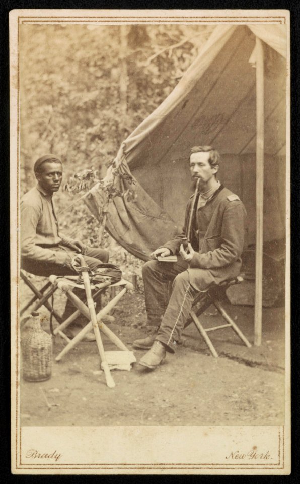 Unidentified soldier of 23rd New York Infantry Regiment smoking a pipe and unidentified African American man, probably a servant, with sword, sitting in front of tent) - Brady, New York LCCN2016652228
