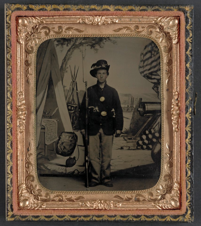 Unidentified soldier of 33rd Missouri Infantry Regiment with bayoneted musket and revolver in front of painted backdrop showing weapons and American flag at Benton Barracks, Saint Louis, LCCN2012646966