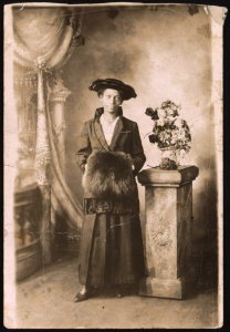 Unidentified woman, posing for studio portrait, wearing hat, coat, and muffler, standing next to a vase of flowers on a pedestal LCCN2015649997