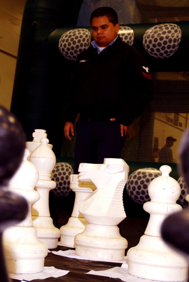 US Navy 070124-N-3038W-088 Electrician's Mate 2nd Class Robert Ramirez ponders his next move on the giant chess board during the Morale Welfare and Recreation events held in the hangar bay of the Nimitz-class aircraft car