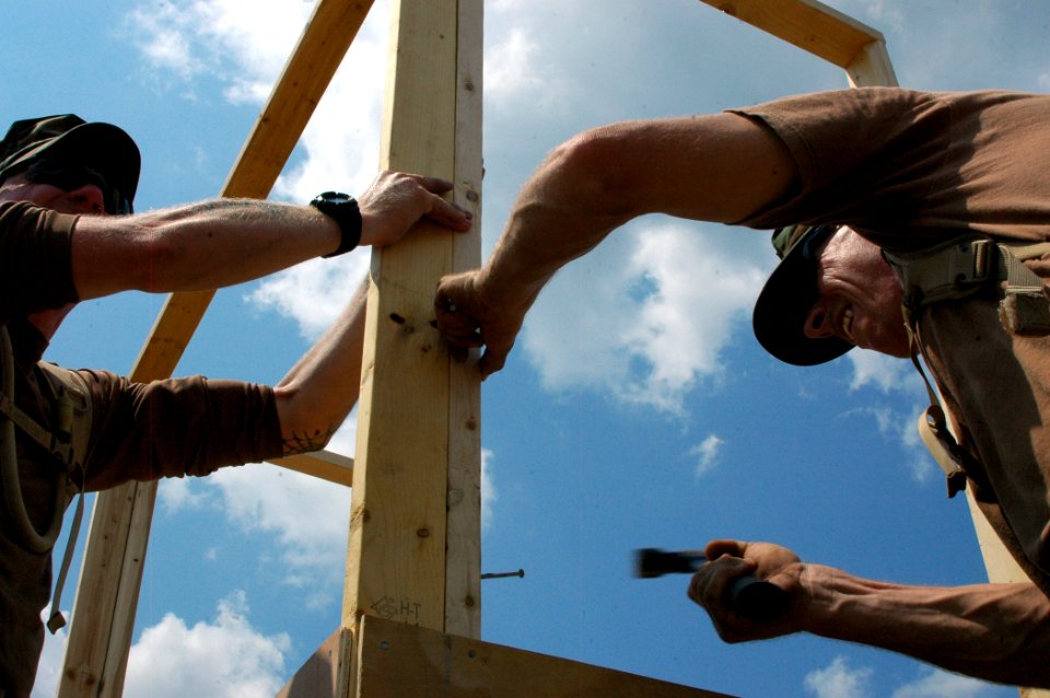 US Navy 070615-N-7367K-001 Steelworker 1st Class David Stephens holds the lumber steady while Builder 1st Class Barry Moyar drives a nail through the structure as they build a command operations center sentry guard shack