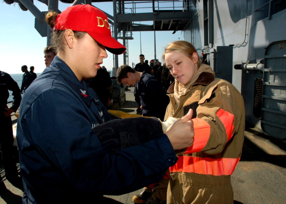 US Navy 071004-N-1655H-001 Information Systems Technician 2nd Class Sylvia Giannetta, a damage control training team member, assists Hospital Corpsmen 3rd Class Patricia Meir with her fire fighting ensemble during the Damage Co