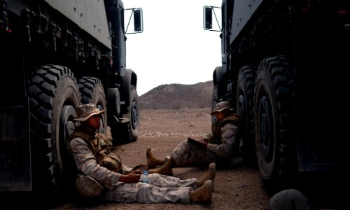 US Navy 080123-F-1644L-101 Cpl. Steven Pepper, left, and Lance Cpl. David Hernandez both assigned to the 3rd Low Altitude Air Defense Battalion, eat a meals, ready-to-eat (MRE) and read a book in between two seven-ton trucks