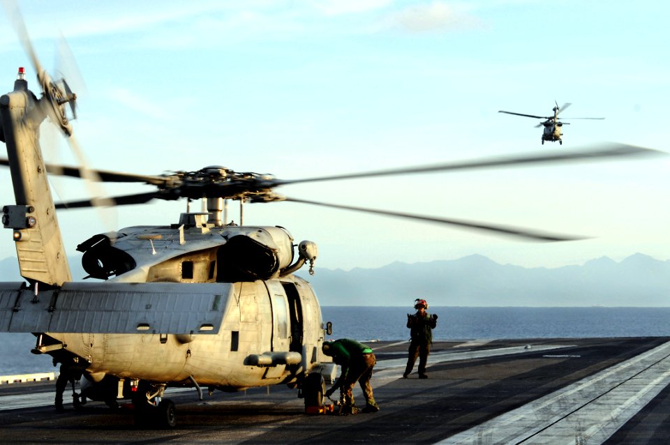 US Navy 080701-N-5961C-004 An SH-60F Seahawk flies off in the distance as crewmen and maintenance crews assigned to Helicopter Anti-Submarine Squadron (HS) 4 prepare a second helicopter for launch from the aircraft carrier USS