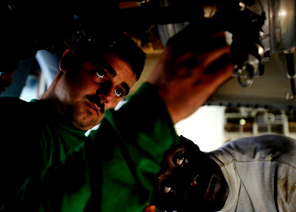 US Navy 081023-N-7571S-002 Aviation Machinist's Mate 2nd Class Prince Agyemang observes Aviation Machinist's Mate 3rd Class Nicholas Costello as he fixes an oil cap lanyard
