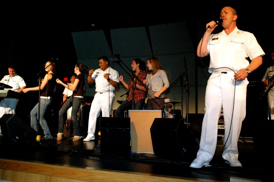 US Navy 090527-N-7975R-006 Musician 1st Class Ira Ostrowski sings lead vocals with the Naval Academy Band, the Electric Brigade, as Chief Musician Frank Dominguez dances with students during a performance at Irondequoit High
