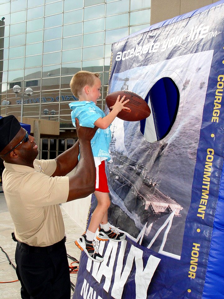 US Navy 090613-N-2888Q-002 Master-at-Arms 1st Class Samuel Armah, left, from Navy Recruiting District Dallas, helps a child deliver a touchdown pass into an inflatable U.S. Navy football toss during an Oklahoma Navy Week event